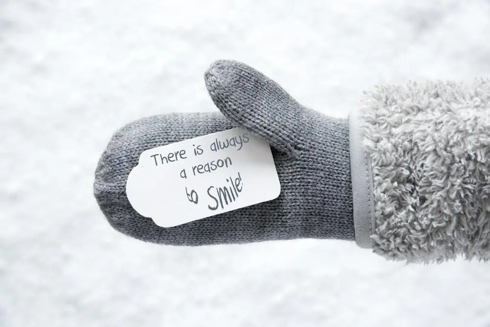 gloved hand in the snow holding a tag that reads "be someones reason to smile"